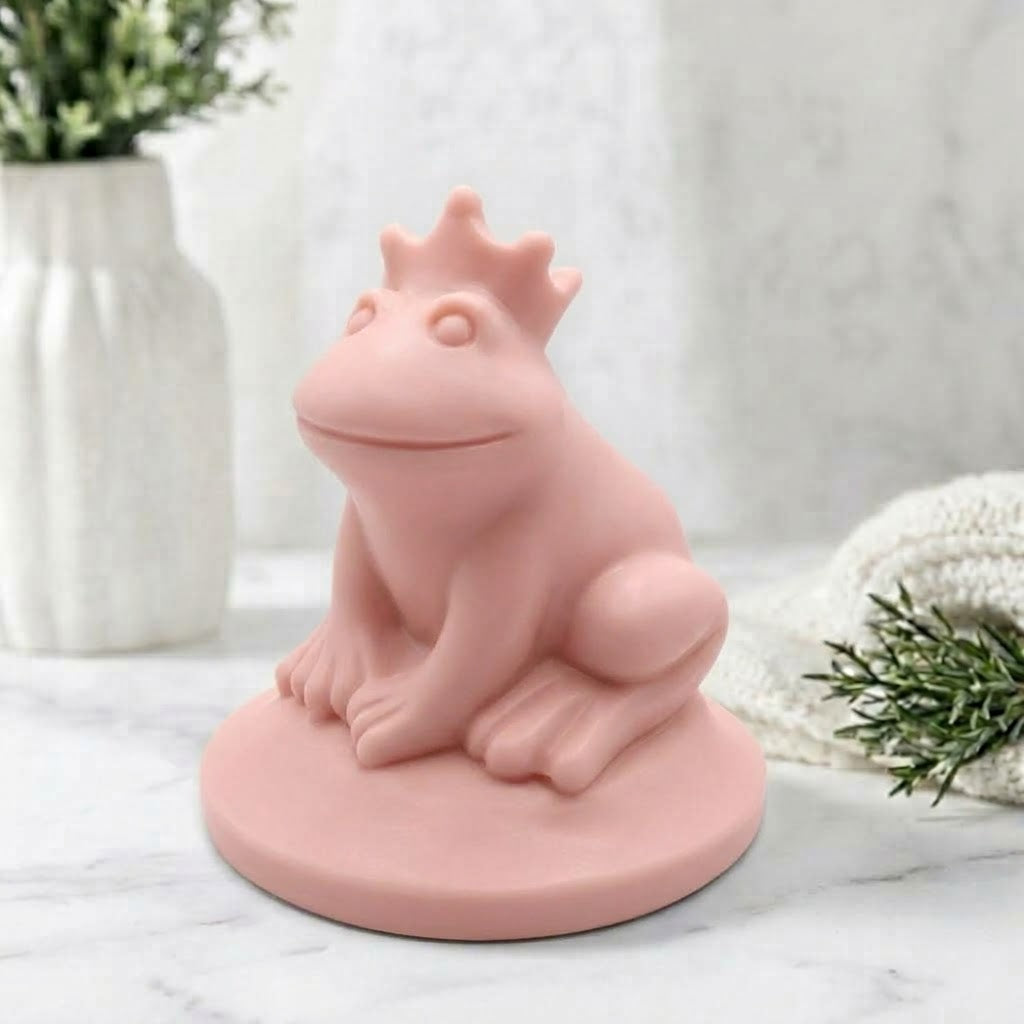 A side view of a soft pink handmade vegan soap featuring a frog prince with a crown on his head, displayed on a white background