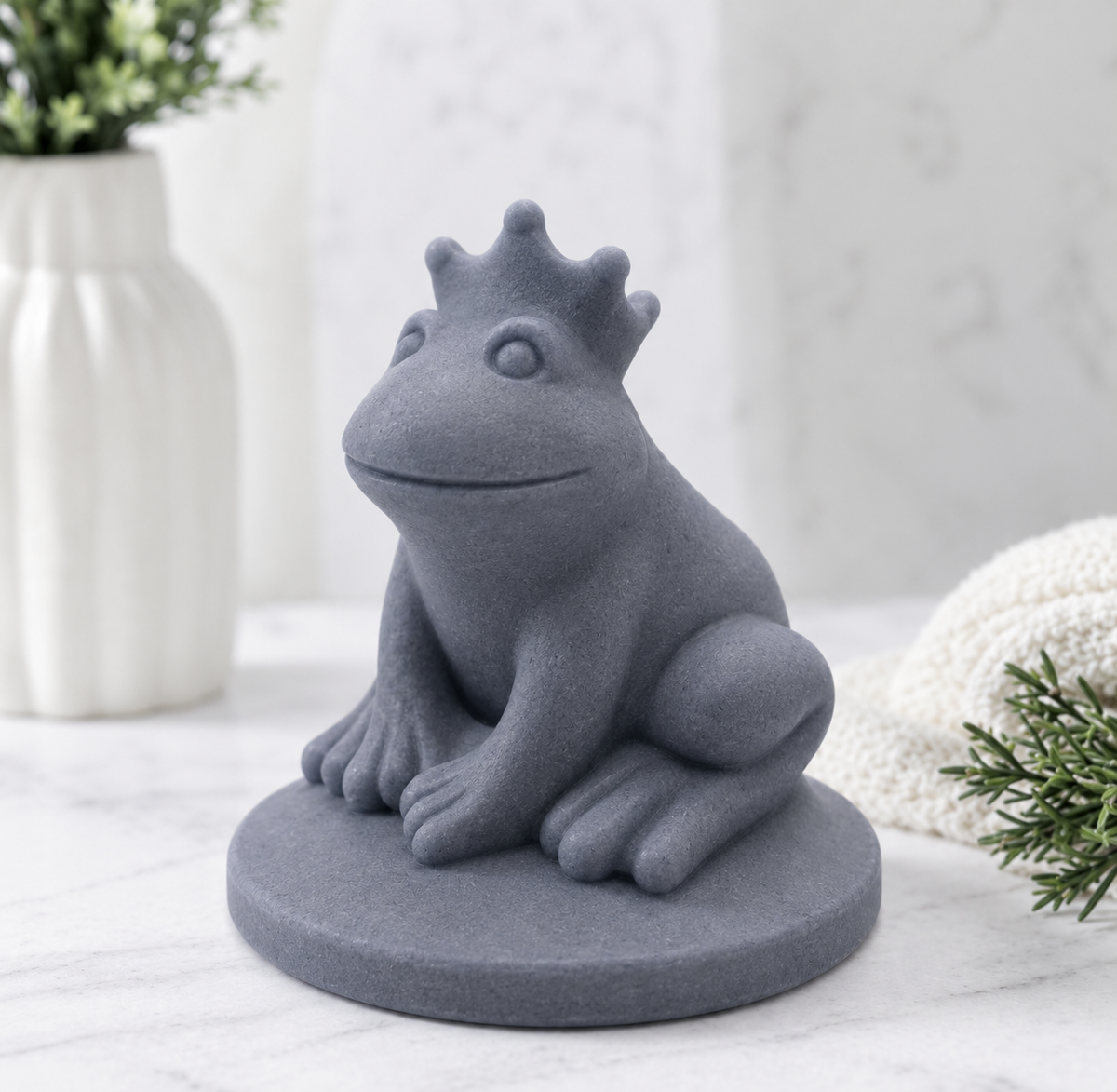 A side view of a charcoal grey handmade vegan soap featuring a frog prince with a crown on his head, displayed on a white background