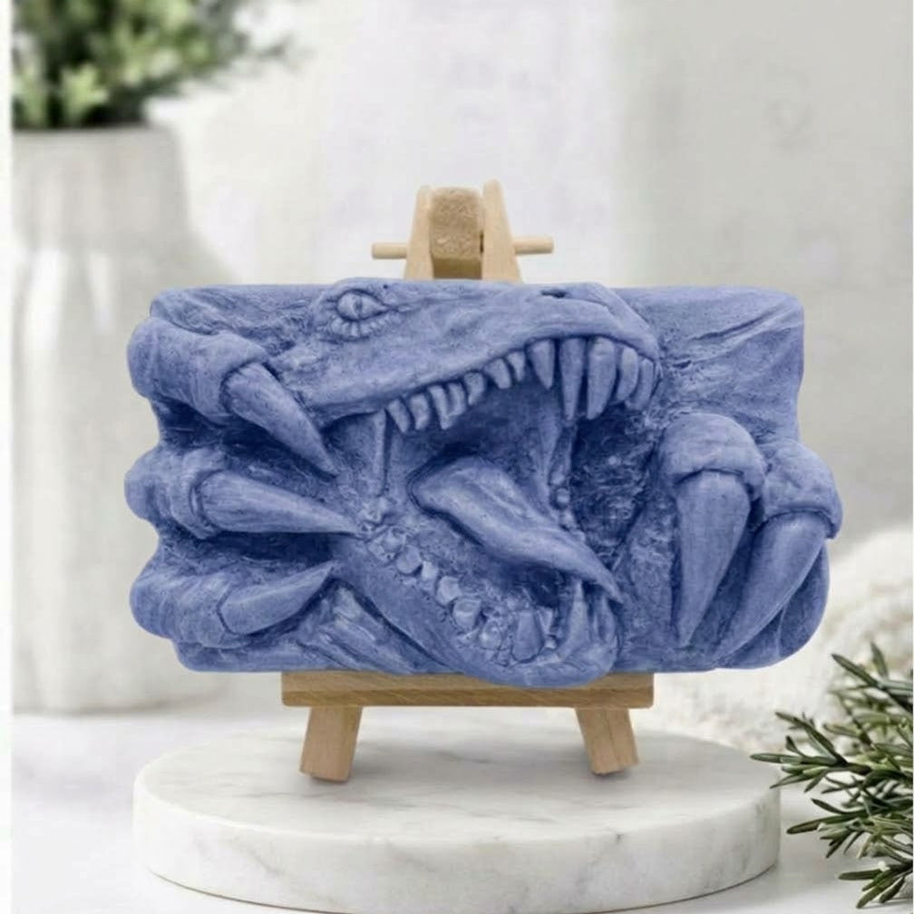 A rectangular soft blue handmade vegan soap  featuring a high-relief carving of a Tyrannosaurus Rex with its jaws wide open in a roar and sharp claws framing the face, displayed on a miniature wooden easel.