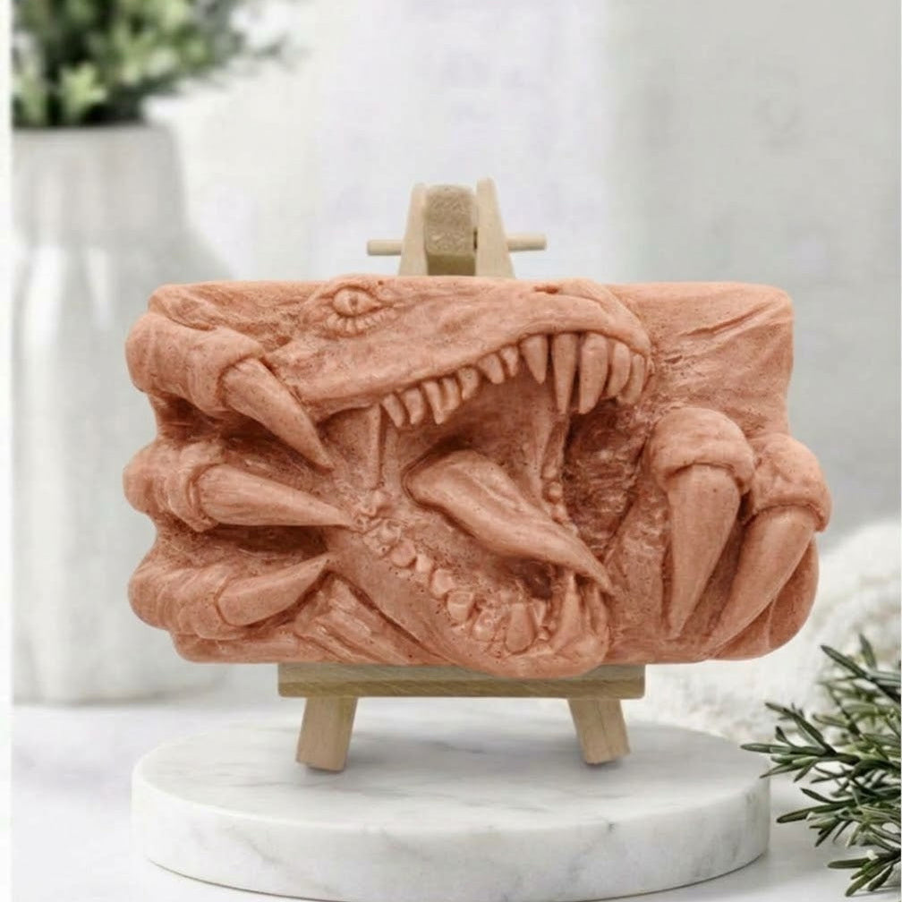 A rectangular terracotta handmade vegan soap  featuring a high-relief carving of a Tyrannosaurus Rex with its jaws wide open in a roar and sharp claws framing the face, displayed on a miniature wooden easel.