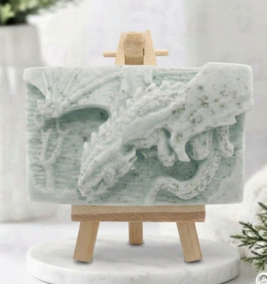 handmade soft green soap bar featuring a detailed relief carving of a dragon, displayed on a small wooden easel in a bright, rustic room with white flowers in the background.