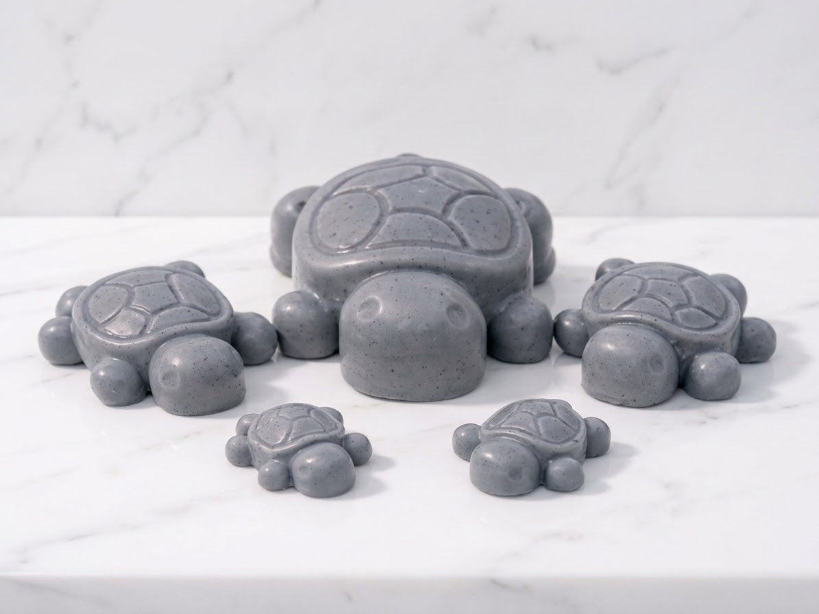 A set of five grey-charcoal coloured artisan turtle soap bars, arranged as a family set (one large, two medium, two small) on a marble countertop. Novelty soap gift for men or kids.