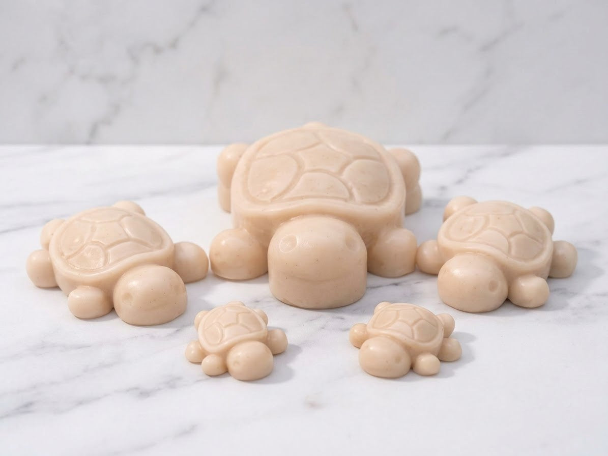 A set of five soft yellow coloured handmade turtle soap bars, arranged in a family grouping of large, medium, and small sizes, displayed on a polished white marble surface. Unique novel soap gift.