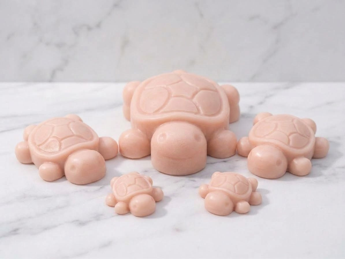 A family grouping of five pale pink-rose coloured handmade turtle soap bars, featuring one large and two smaller pairs, displayed on a polished white marble surface. Romantic or unique spa gift idea.