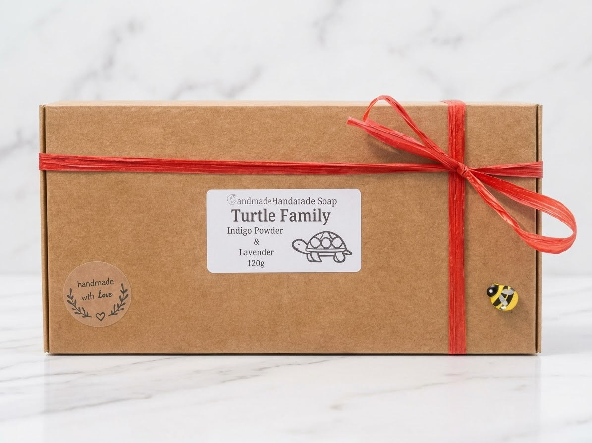 A sustainable brown kraft cardboard soap gift box, tied with a red raffia ribbon and a bow, featuring a specific label for "Turtle Family" handmade soaps scented with Indigo Powder and Lavender. Decorative "handmade with Love" stickers and a bee charm. Unique artisan soap packaging.