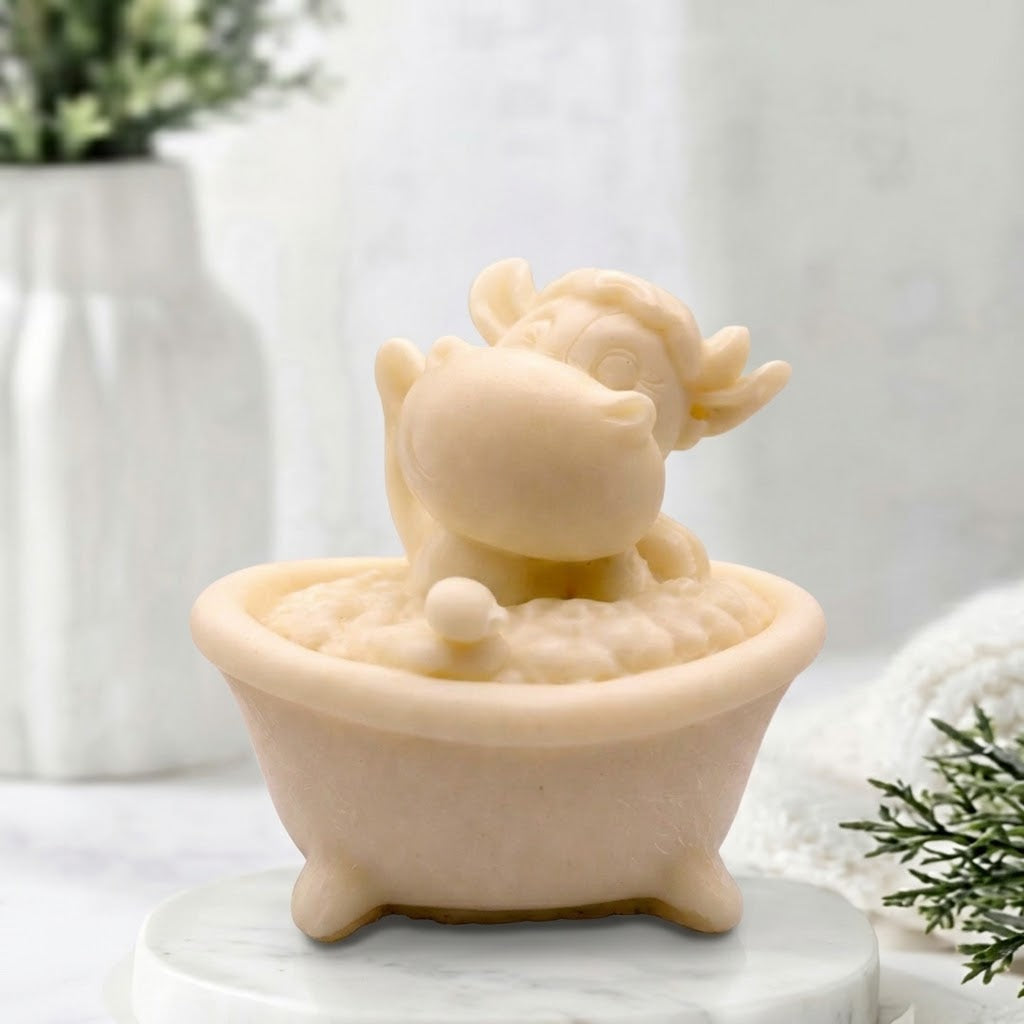 Novelty vegan bar soap of a cute soft yellow cartoon cow figure happily waving while sitting inside a miniature footed bathtub filled with soap bubbles.