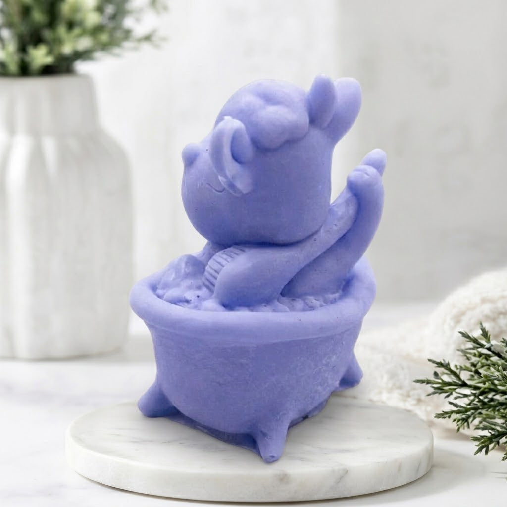 Side view of a novelty handmade vegan soap bar in lavender blue. Cartoon cow figure sitting within a miniature bathtub structure.