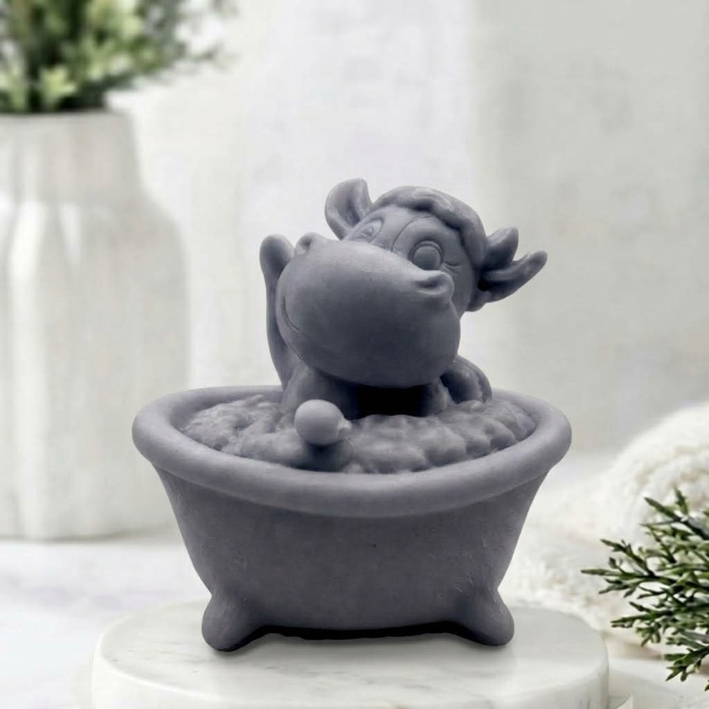 Cute animal novelty vegan soap bar sculpted as a dark grey charcoal cartoon cow happily sitting inside a matching miniature footed bathtub surrounded by bubbles.