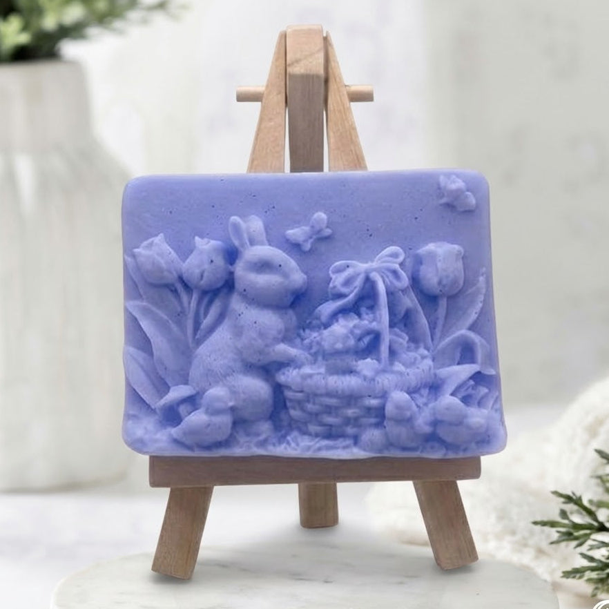 Artisanal lavender blue vegan soap bar with a detailed raised carving of an Easter bunny next to a flower-filled basket, displayed on a small wooden easel.