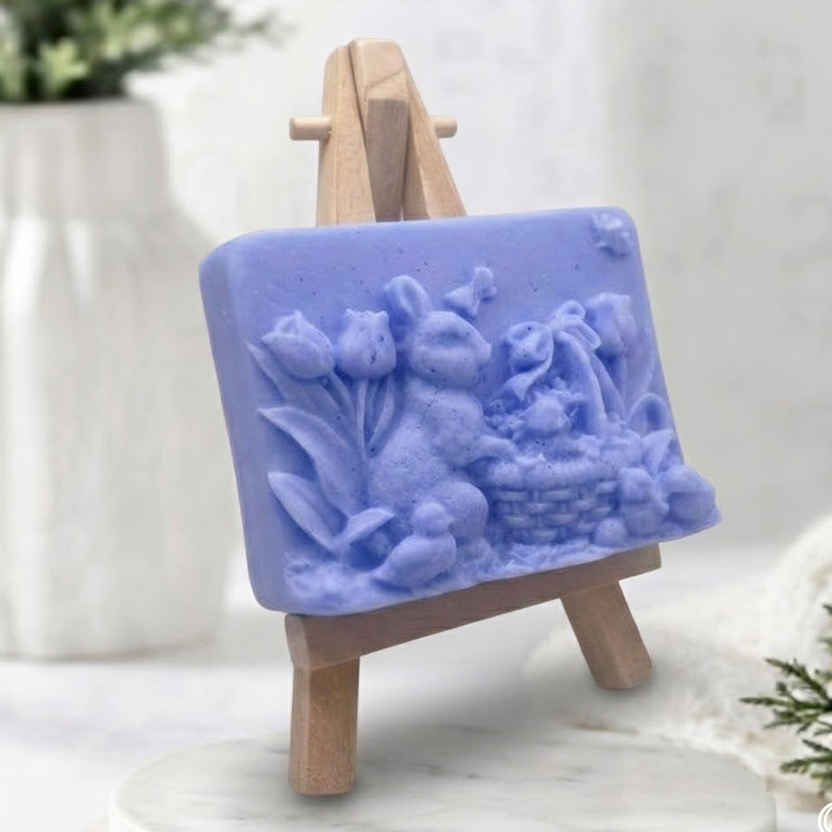 An angled view of a handcrafted light blue vegan Easter soap featuring a sculpted relief design of a rabbit, spring tulips, and a basket on a mini wooden display stand.