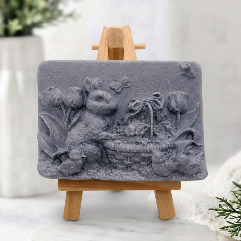 Sculpted charcoal grey vegan soap with a detailed carving of an Easter rabbit, eggs, and spring flowers, presented on a mini wooden easel.