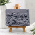 Sculpted charcoal grey vegan soap with a detailed carving of an Easter rabbit, eggs, and spring flowers, presented on a mini wooden easel.