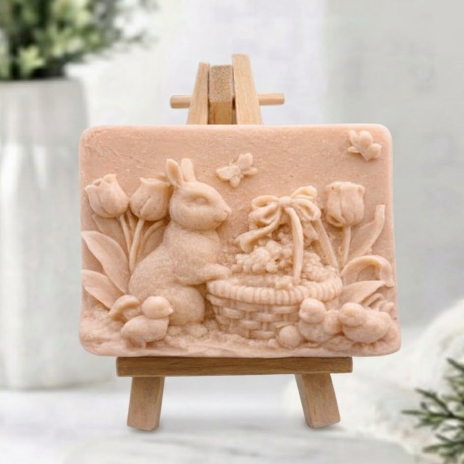 Pink coloured handmade vegan soap bar with an intricate raised design of an Easter bunny, a woven basket, and tulips on a small wooden tripod easel.