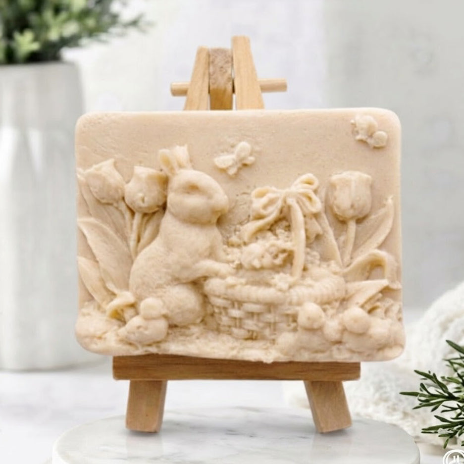 Artisan soft yellow coloured vegan soap bar featuring a detailed sculpted relief of an Easter rabbit with a basket of eggs and spring flowers on a mini wooden display easel.