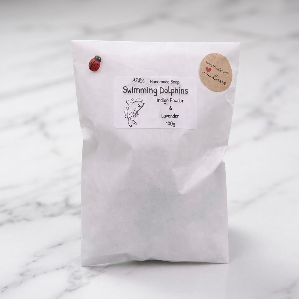 Eco-friendly white paper pouch packaging for the 'Swimming Dolphins' handmade vegan soap, featuring a ladybug pin, a 'handmade with love' sticker, and a label specifying Indigo Powder & Lavender ingredients, 100g.