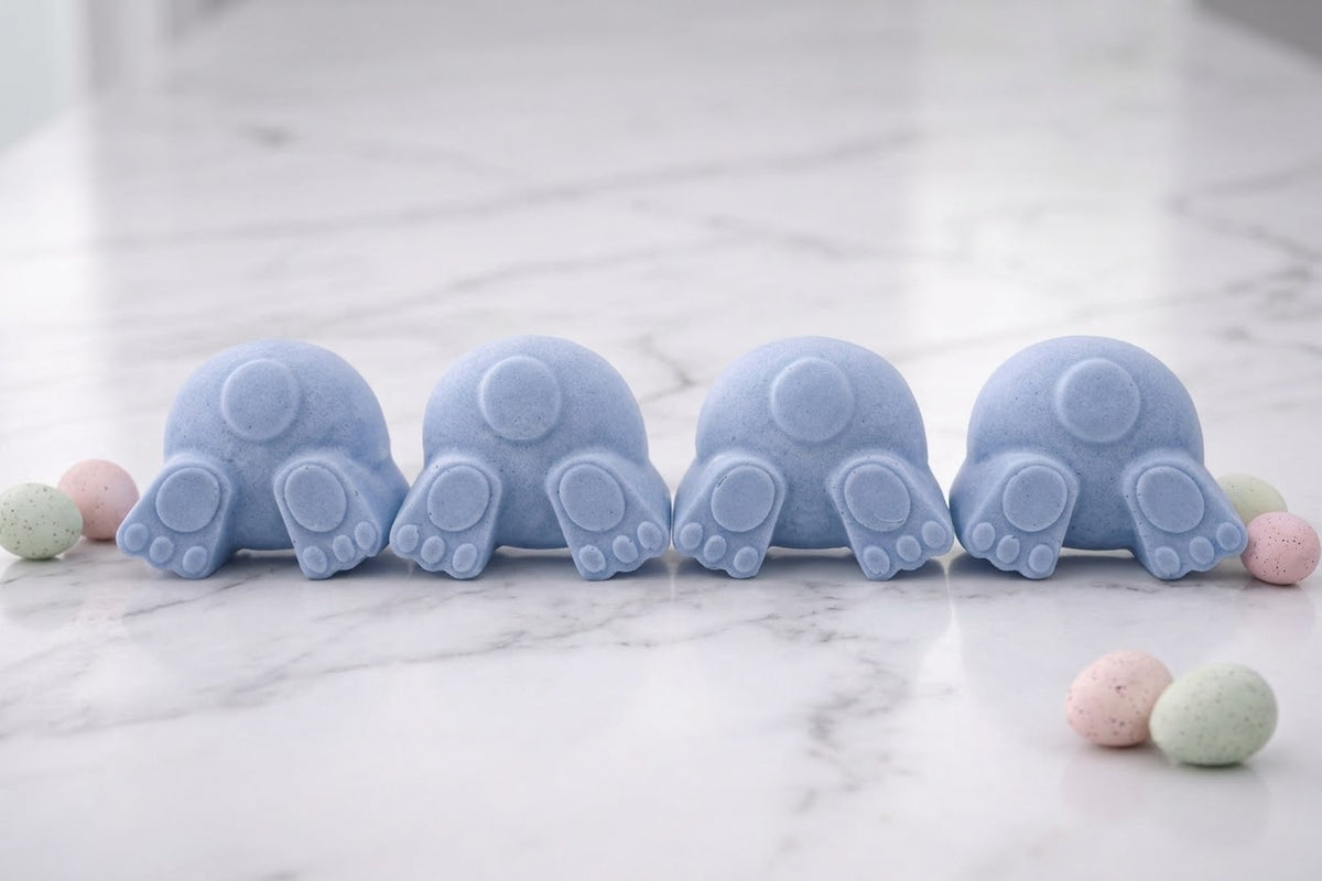 Set of four pastel blue vegan soap bars shaped like bunny bottoms with tails, arranged horizontally on a white marble background with decorative Easter eggs.
