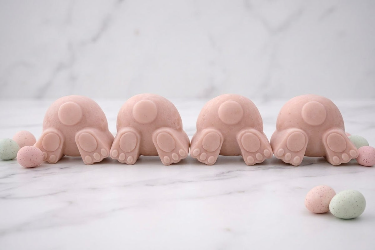 Handmade soft pink vegan soap bunnies displaying their tails and paw prints. A collection of four novelty soaps on a marble countertop with speckled holiday eggs.