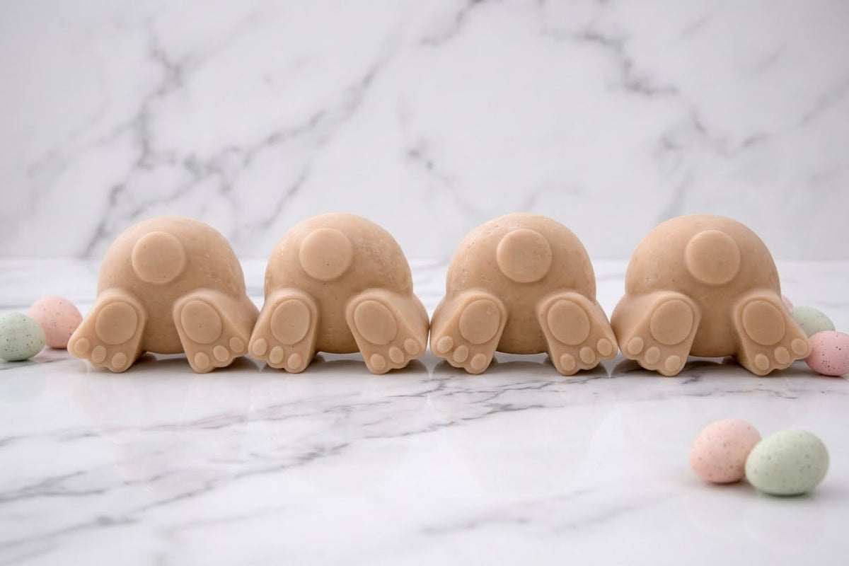 Collection of four terracotta coloured vegan soap bunnies showing off their fluffy tails and paw pads. Arranged on a clean marble table with scattered pastel speckled Easter egg decorations. A charming Easter gift idea.