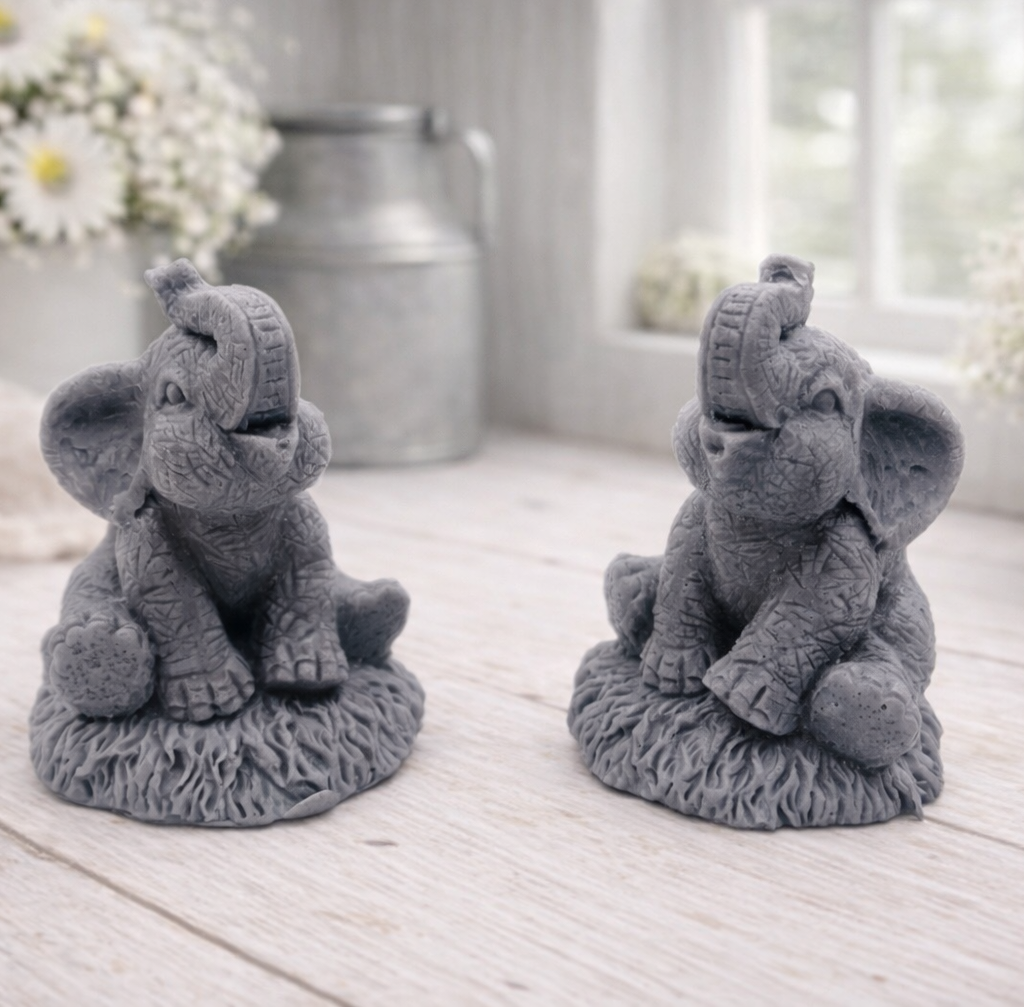 A charming gift set of two handmade vegan soaps, one sculpted as a sitting baby elephant with a trunk raised in a greeting and the other as a smaller standing calf, displayed on a light wooden surface in a bright farmhouse setting.