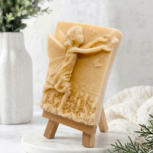 Side view of a soft yellow coloured, relief-carved vegan fairy soap bar with a handcrafted artisan fairy and butterfly pattern, standing on a miniature wooden easel. Natural essential oil soap, self-care gift.