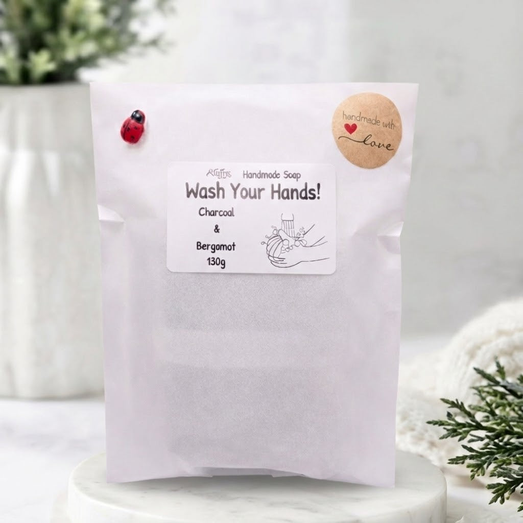 paper pouch packaging for handmade charcoal and bergamot vegan soap, labeled "Wash Your Hands!", featuring a ladybug sticker and "handmade with love" label, displayed on a marble stand with blurry spa decor background.