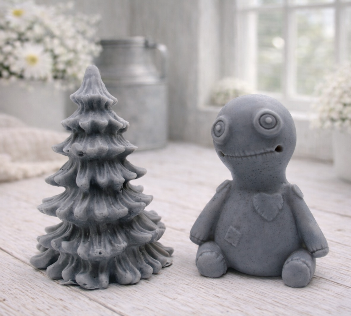 A handmade vegan soap gift set featuring a cream-coloured voodoo doll soap with stitched details and a grey tiered Christmas tree soap, presented in a black gift box tied with a red raffia ribbon and a small ladybird embellishment.