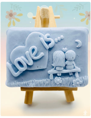 Handmade vegan soaps featuring a boy and girl sat on a bench with the wording Love Is... ,  in beautiful eco friendly packaging. crafted with natural French clays, activated charcoal and essential oils. a eco-friendly, zero waste gift that’s cruelty-free, skin-nourishing, the perfect gift for Valentines day or anniversary gift. small business UK