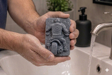Load image into Gallery viewer, Person holding a bar of soap with decorative detailed handmade vegan soap featuring a motorcycle rider with a skull emblem on their jacket, displayed on a wooden easel against a whimsical farmhouse decor background
