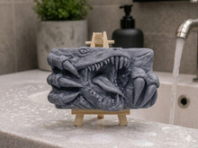 Load image into Gallery viewer, A rectangular handmade vegan soap in a charcoal-grey tone, featuring a high-relief carving of a Tyrannosaurus Rex with its jaws wide open in a roar and sharp claws framing the face, displayed on a miniature wooden easel.

