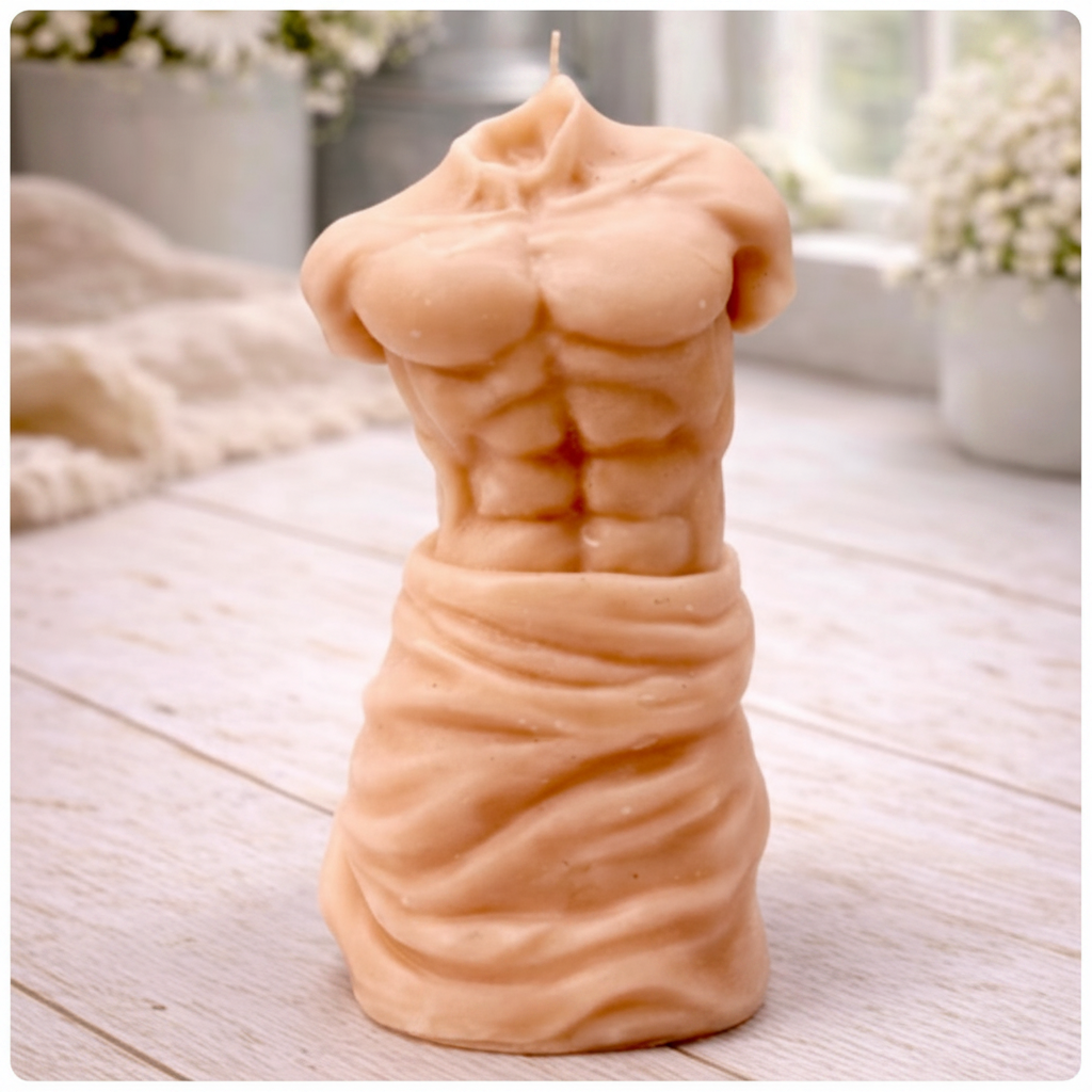 A handmade vegan soap sculpted into a realistic male torso with defined abdominal muscles and draped fabric at the waist, in a natural flesh tone, set against a bright window and floral background.