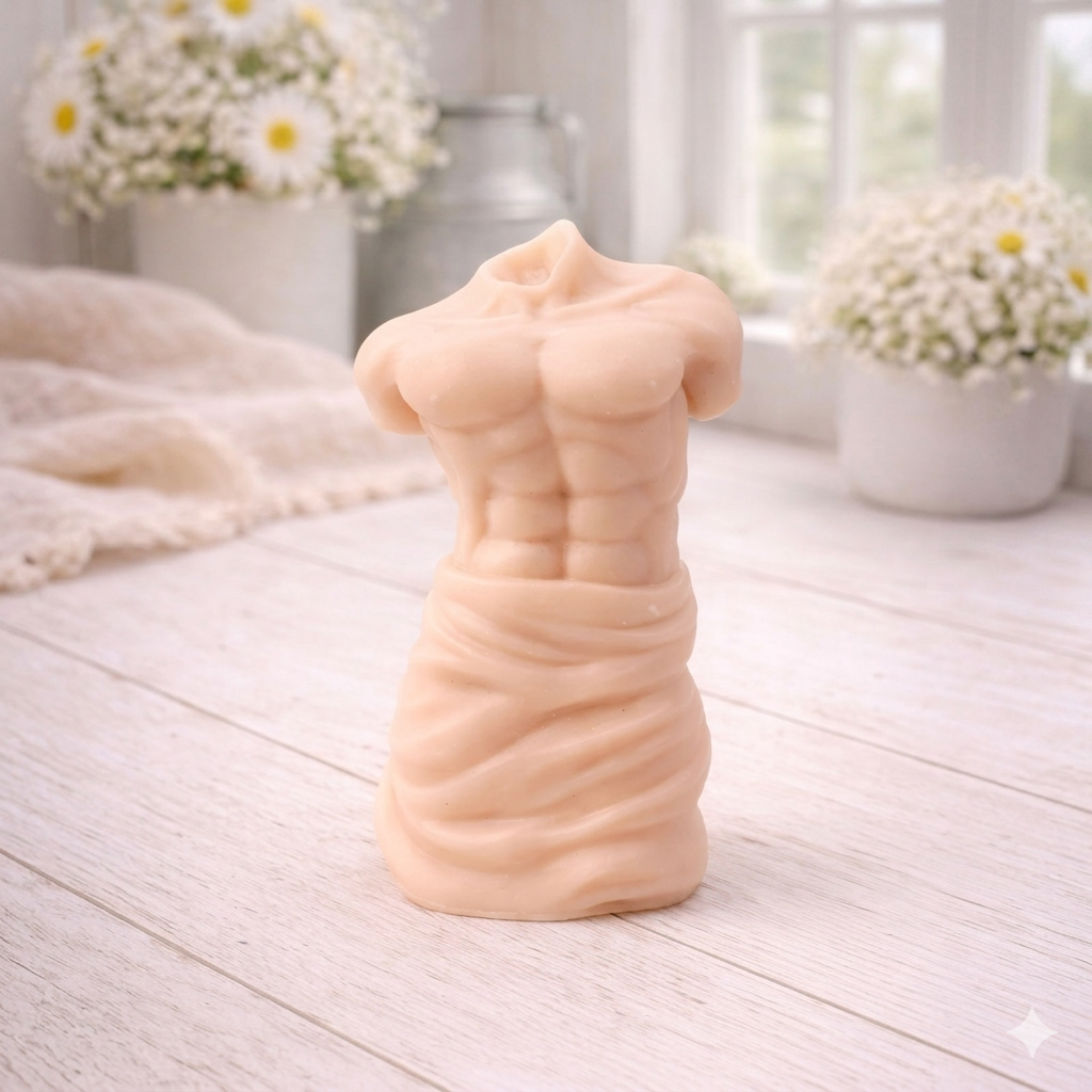 A handmade vegan soap sculpted into a realistic male torso with defined abdominal muscles and draped fabric at the waist, in a natural flesh tone, set against a bright window and floral background.