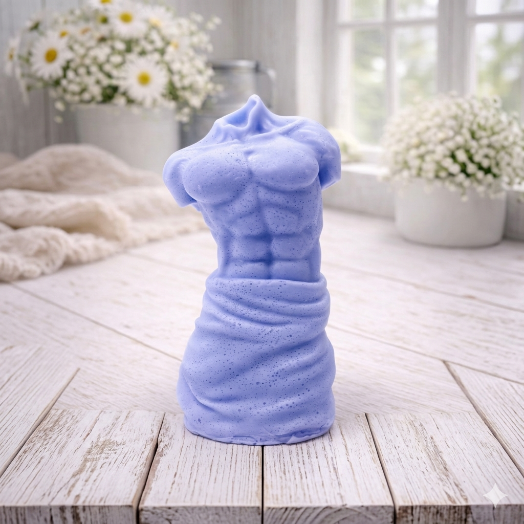 A handmade vegan soap sculpted into a realistic male torso with defined abdominal muscles and draped fabric at the waist, in a natural flesh tone, set against a bright window and floral background.