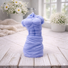 Load image into Gallery viewer, A handmade vegan soap sculpted into a realistic male torso with defined abdominal muscles and draped fabric at the waist, in a natural flesh tone, set against a bright window and floral background.
