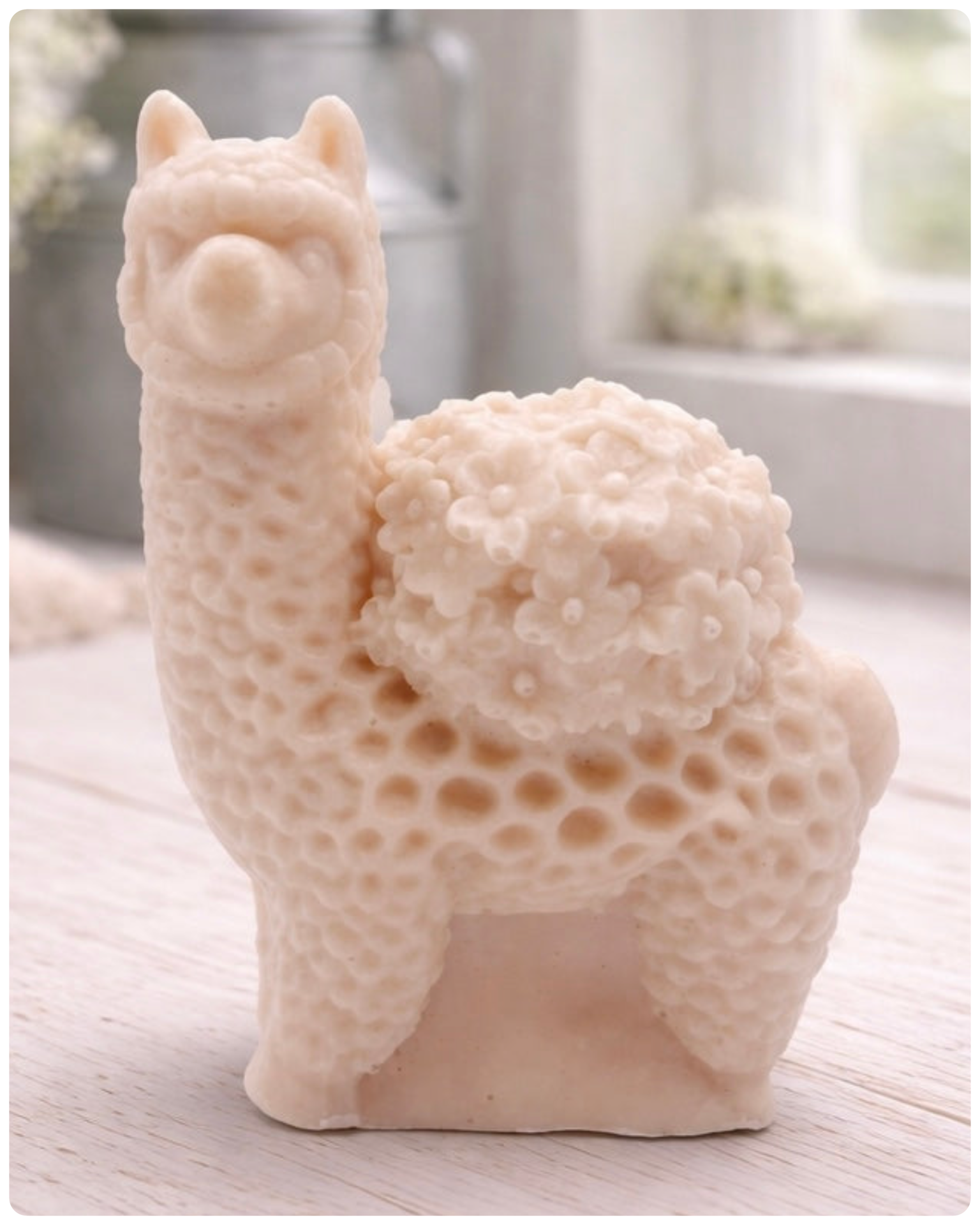A handmade vegan soap featuring a charming llama or alpaca with a highly textured, fleecy coat and a blanket of intricate flowers across its back, displayed on a light wooden surface in a bright room.