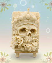 Load image into Gallery viewer, A handmade vegan soap featuring a highly detailed 3D relief of a skull wearing a jewelled crown and surrounded by roses, displayed on a wooden easel against a whimsical background.
