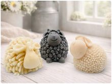 Load image into Gallery viewer, A set of three handmade vegan soaps in different sheep styles, including a dark grey smiling sheep and two lambs with detailed woolly textures, displayed on a rustic wooden surface with lavender and flowers.
