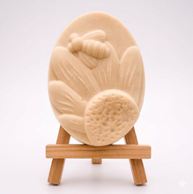 Load image into Gallery viewer, A handmade vegan soap in a natural colour, featuring an oval relief of a honeybee landing on a large sunflower with detailed petals and a textured centre, displayed on a small wooden easel.
