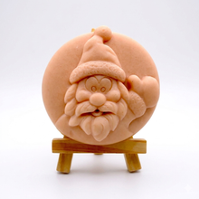 Load image into Gallery viewer, A circular handmade vegan soap featuring a detailed high-relief carving of a jolly Father Christmas waving, with a textured beard and a classic Santa hat, displayed on a miniature wooden easel.
