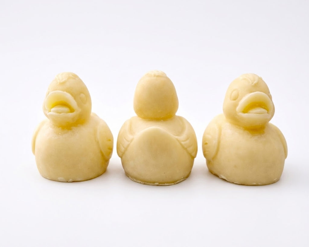 Three handmade vegan soaps in the shape of fluffy ducklings with open beaks, displayed on a neutral linen cloth in a bright farmhouse decor setting with Easter eggs in the background.