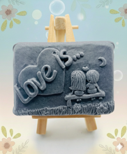 Load image into Gallery viewer, Handmade vegan soaps featuring a boy and girl sat on a bench with the wording Love Is... ,  in beautiful eco friendly packaging. crafted with natural French clays, activated charcoal and essential oils. a eco-friendly, zero waste gift that’s cruelty-free, skin-nourishing, the perfect gift for Valentines day or anniversary gift. small business UK
