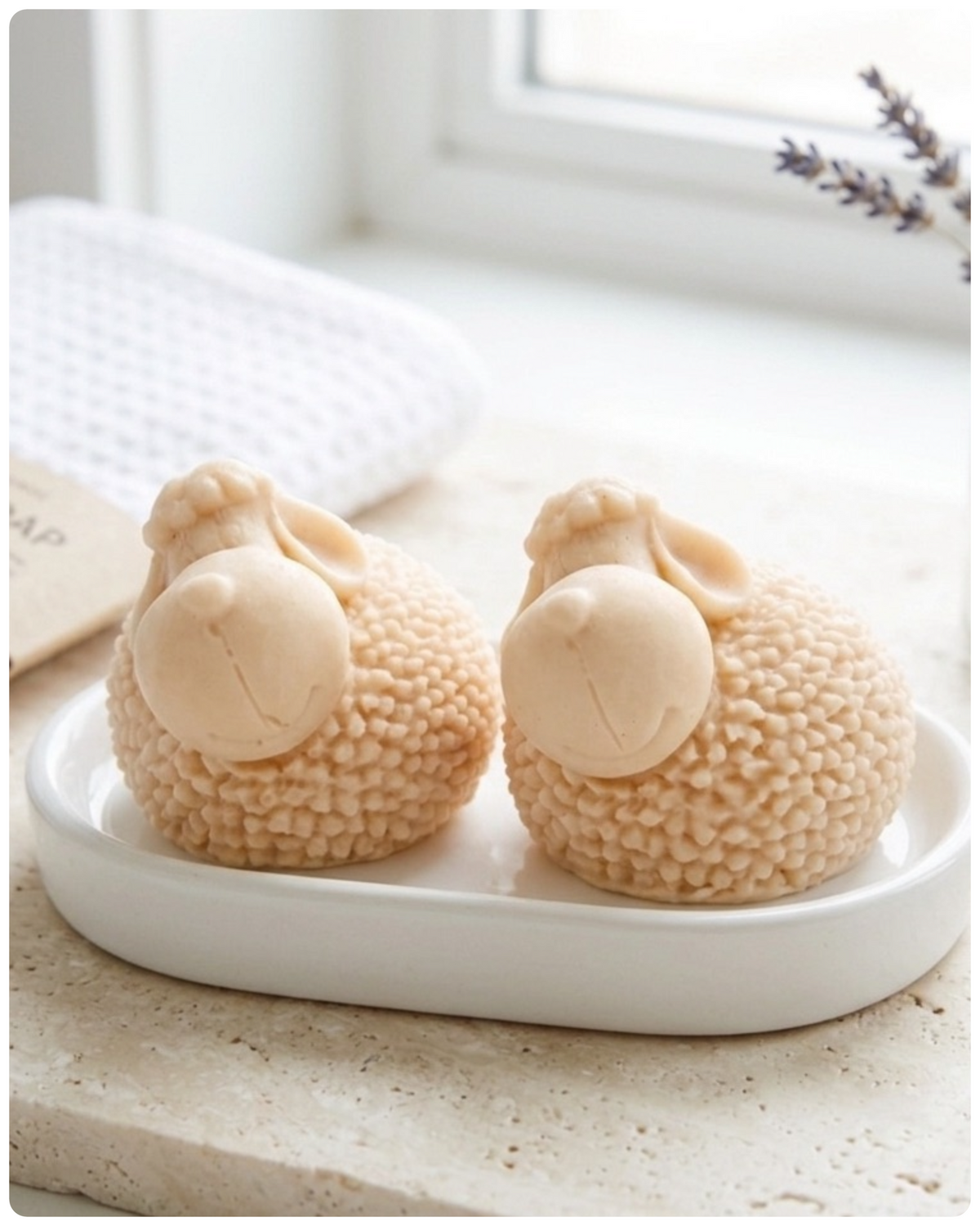 Two handmade vegan soaps in the shape of fluffy spring lambs with detailed wool texture, displayed on a weathered white wooden surface in a bright farmhouse decor room