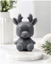 Load image into Gallery viewer, A 3D handmade vegan soap crafted into a cute, sitting baby reindeer with small antlers, rounded features, and a sweet expression, set against a bright farmhouse window.
