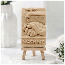 Load image into Gallery viewer, handmade vegan soap featuring a relief sculpture of hands being washed under a dripping tap with the words "WASH ME" at the base, displayed on a small wooden easel in a bright farmhouse decor kitchen setting.
