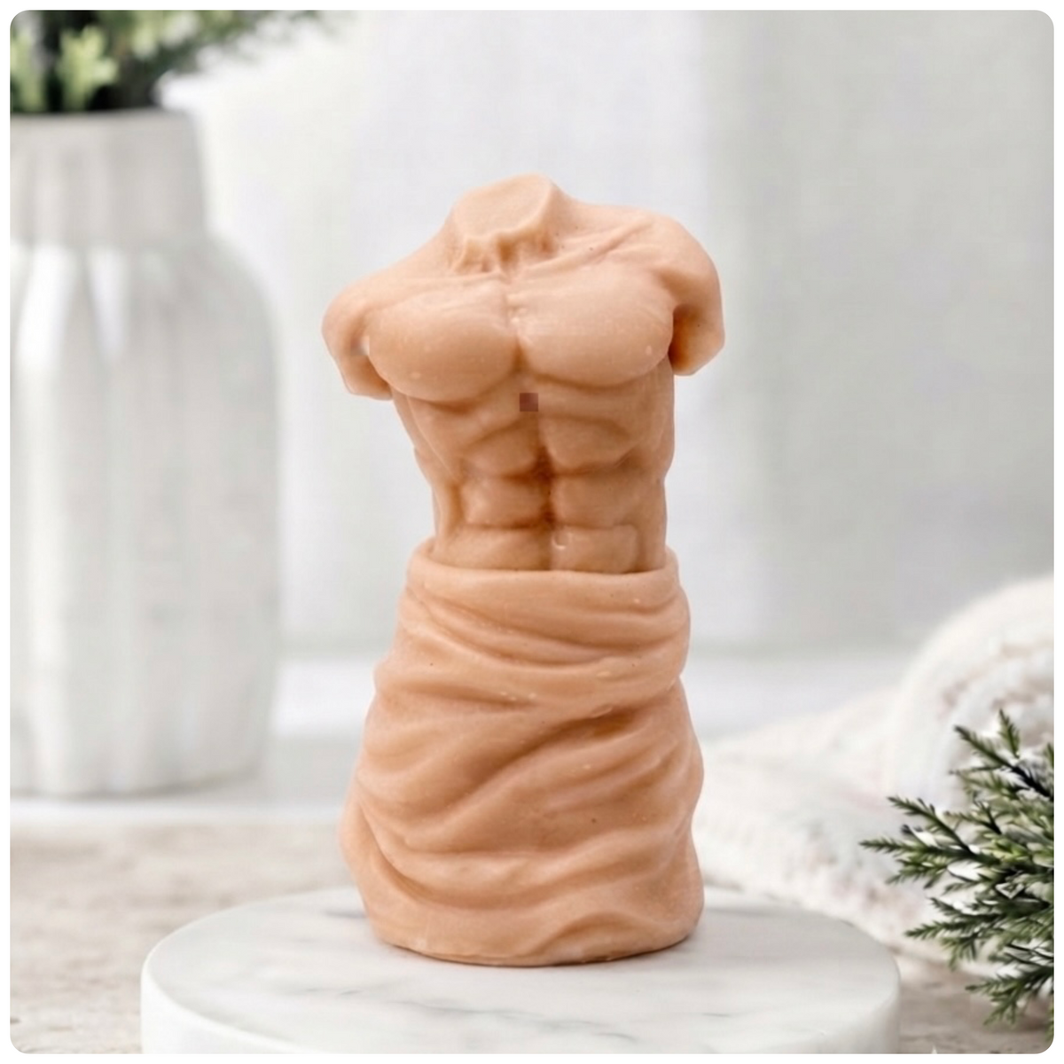 A handmade vegan soap sculpted into a realistic male torso with defined abdominal muscles and draped fabric at the waist, in a natural flesh tone, set against a bright window and floral background.