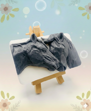 Load image into Gallery viewer, A grey and white handmade vegan soap featuring a highly detailed relief of two horses nuzzling heads, displayed on a wooden easel against a whimsical farmhouse decor background.
