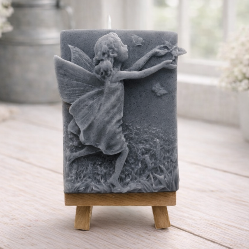 A rectangular handmade vegan soap featuring a detailed relief of a winged fairy reaching towards a butterfly in a meadow of tall grass and flowers, displayed on a miniature wooden easel