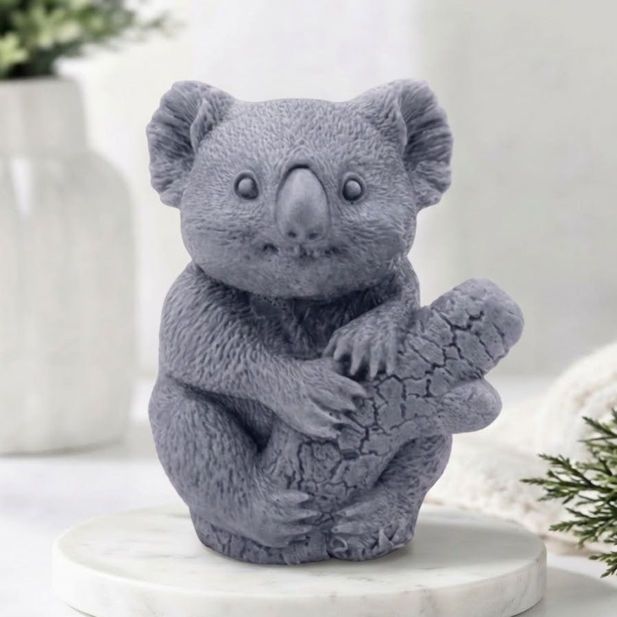 A front-facing photograph of a grey, charcoal-coloured koala-shaped soap bar. The highly detailed soap is sculpted to show textured fur and the koala clinging to a textured log. It sits on a round, white marble disc. In the blurry background, there is a white ceramic vase with green sprigs and a rolled white towel.