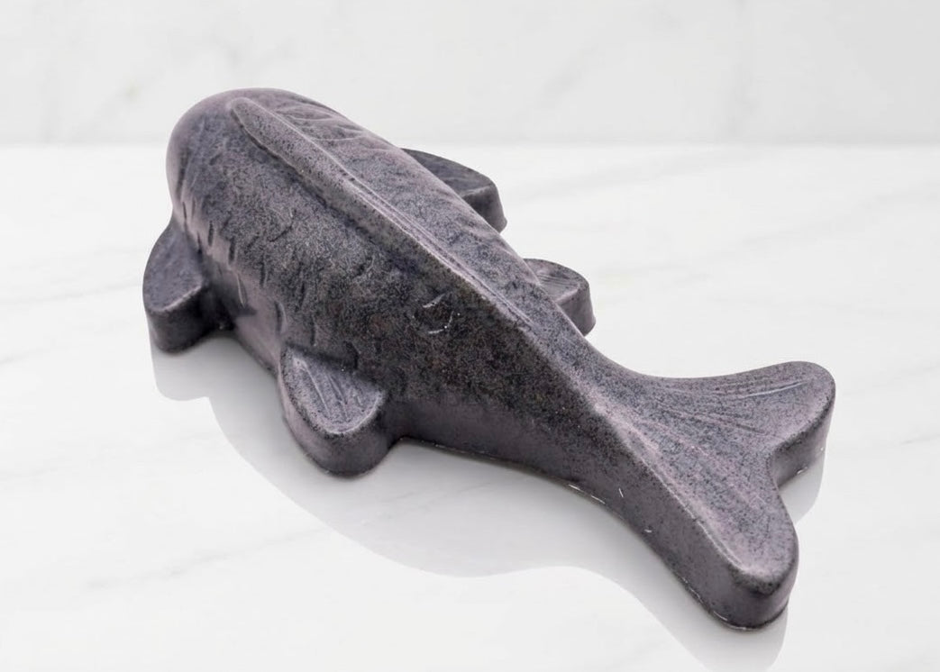 Handmade charcoal vegan soap sculpted in the shape of a koi carp fish with detailed scales and fins, resting on a light timber surface with a soft floral background.