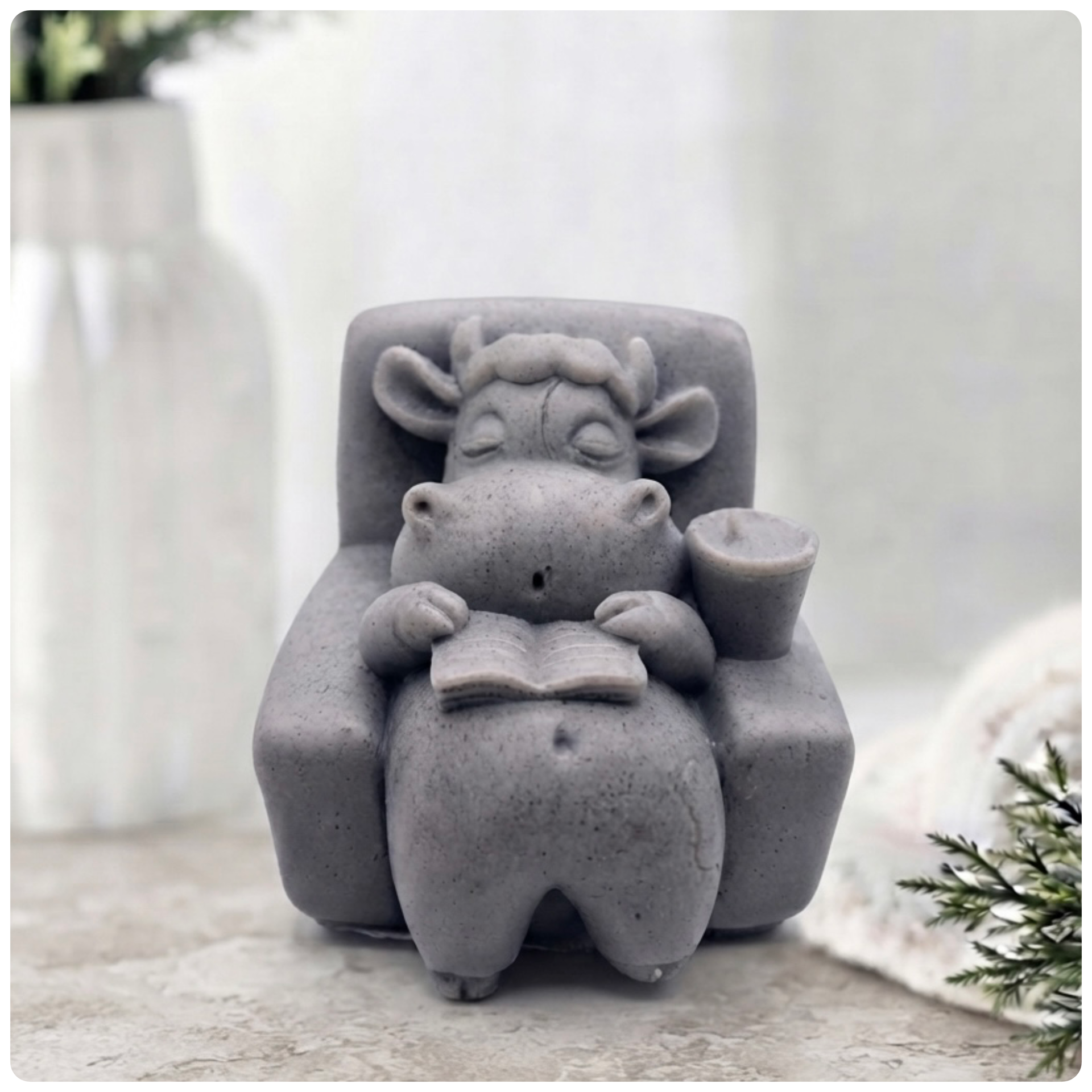 A handmade vegan soap sculpted to show a whimsical "lazy cow" character relaxing in an armchair, reading a book and holding a drink, displayed on a light wooden surface.