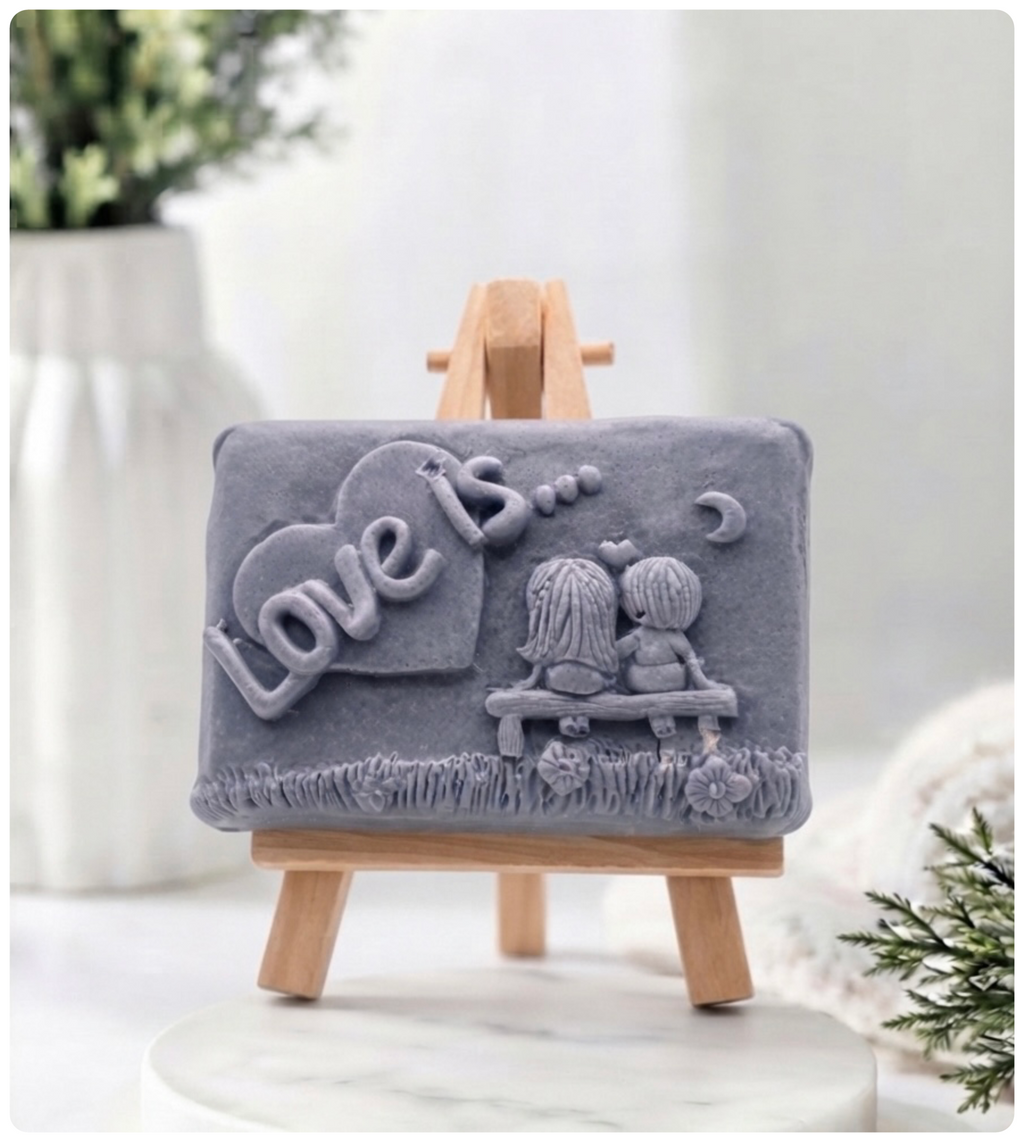 A rectangular handmade vegan soap featuring a detailed 3D relief carving of a young couple sitting on a bench under a crescent moon, with the words "Love is..." embossed inside a heart, displayed on a wooden easel.